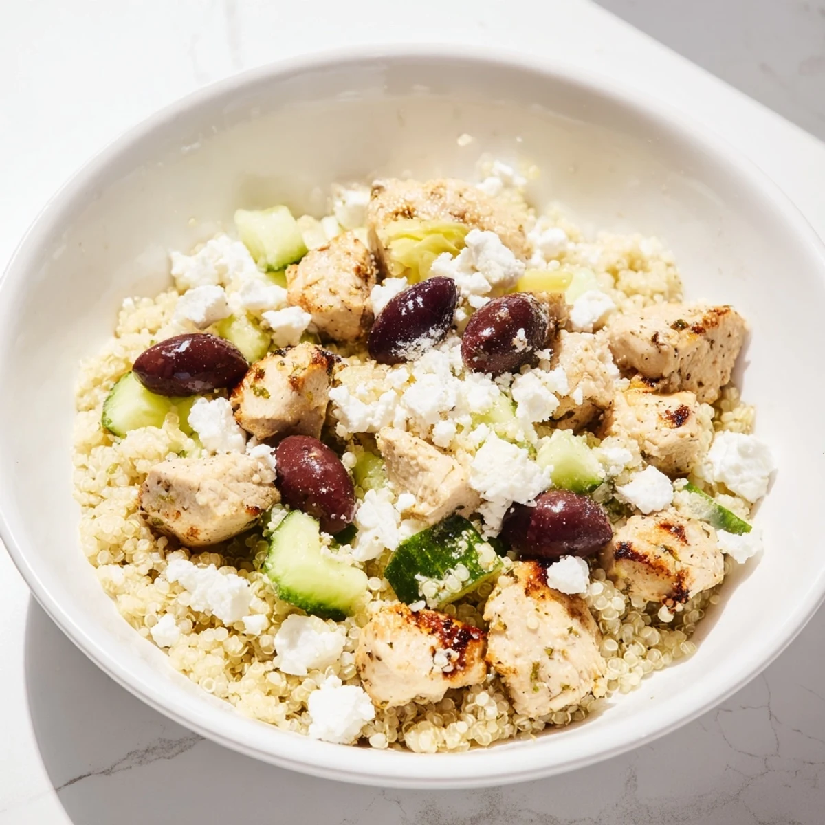 Photo shows Mediterranean Chicken Bowl served in a white bowl, featuring juicy chicken pieces, chickpeas, feta, olives, and fresh veggies over quinoa, with dressing being poured.