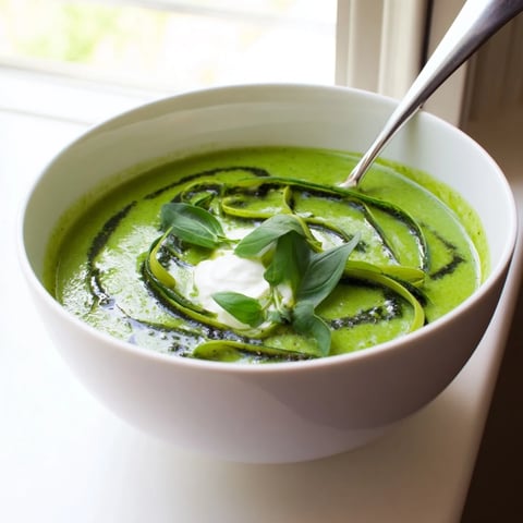 A bowl of vibrant Courgette, Pea and Pesto Soup garnished with fresh basil and a swirl of creamy sauce.