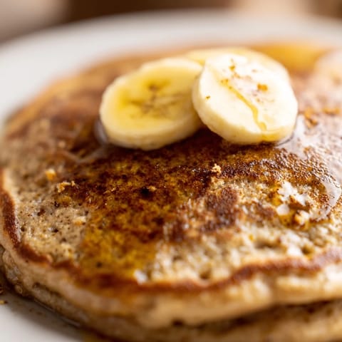 Stack of golden-brown Banana Oat Pancakes topped with sliced bananas, walnuts, and a drizzle of maple syrup on a white plate.