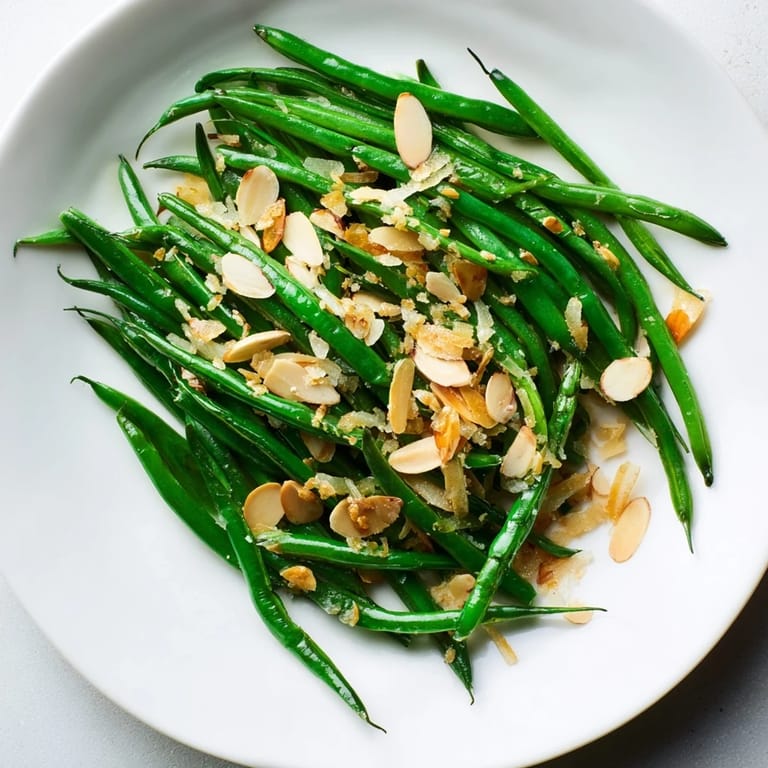 A close-up of tender Sautéed green beans, flavorful garlic and crunchy almonds alongside a dinner plate.