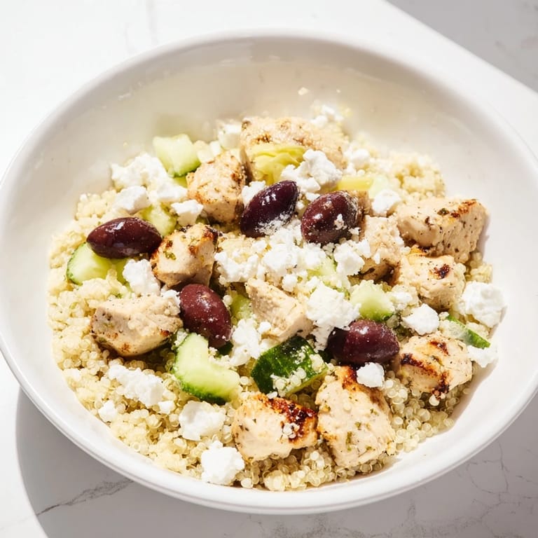Photo shows Mediterranean Chicken Bowl served in a white bowl, featuring juicy chicken pieces, chickpeas, feta, olives, and fresh veggies over quinoa, with dressing being poured.